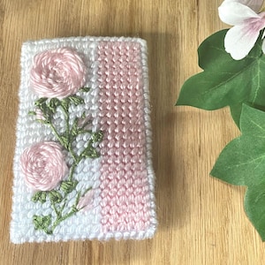 May include: A white and pink knitted case with embroidered pink roses and green stems. The case has a vertical pink stripe and is on a wooden surface. A flower and leaves are in the background.
