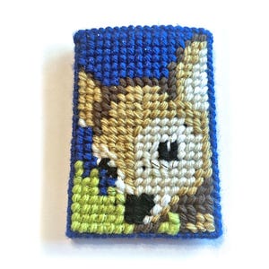 May include: A handmade, rectangular needlepoint depicting a cartoon dog's face. The dog has tan, brown, and white fur, with black eyes and nose. The background is blue, and the grass is green. The edges are also blue.