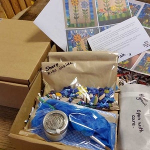 K&ouml;nnte beinhalten: Ein brauner Karton, gef&uuml;llt mit Mosaik-Bastelzubeh&ouml;r. Die Box enth&auml;lt eine T&uuml;te mit bunten Mosaikfliesen, ein kleines Glas mit wei&szlig;em Fugenm&ouml;rtel, ein blaues Tuch und einen Zettel mit der Aufschrift "Scharfe Teile im Inneren".