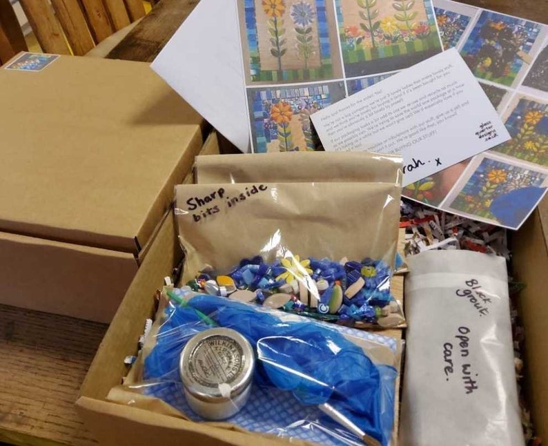 K&ouml;nnte beinhalten: Ein brauner Karton, gef&uuml;llt mit Mosaik-Bastelzubeh&ouml;r. Die Box enth&auml;lt eine T&uuml;te mit bunten Mosaikfliesen, ein kleines Glas mit wei&szlig;em Fugenm&ouml;rtel, ein blaues Tuch und einen Zettel mit der Aufschrift "Scharfe Teile im Inneren".