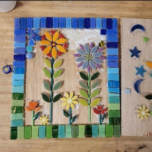 K&ouml;nnte beinhalten: Ein Mosaik-Kunstwerk, das eine Gartenszene mit drei Blumen darstellt. Die Blumen sind aus bunten Keramikfliesen gefertigt und auf einem Holzgrund angeordnet. Die Blumen sind von einem Rand aus blauen und gr&uuml;nen Fliesen umgeben.