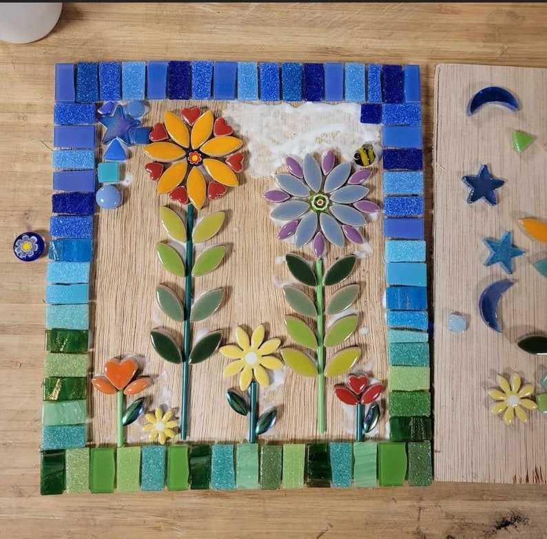 K&ouml;nnte beinhalten: Ein Mosaik-Kunstwerk, das eine Gartenszene mit drei Blumen darstellt. Die Blumen sind aus bunten Keramikfliesen gefertigt und auf einem Holzgrund angeordnet. Die Blumen sind von einem Rand aus blauen und gr&uuml;nen Fliesen umgeben.