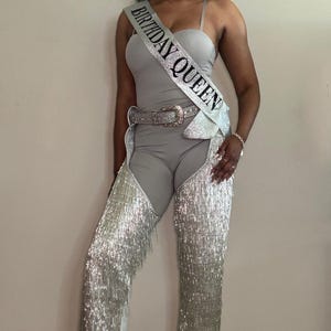 May include: A silver jumpsuit with fringe detailing and a silver belt. The jumpsuit has a scoop neck and a fitted bodice. The person is wearing a silver sash that says "Birthday Queen".