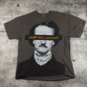 May include: Dark gray t-shirt featuring a black and white portrait of a man with a mustache. A yellow rectangle with the text "CAMP KILL YOURSELF" covers the man's eyes. The shirt is made of a soft material.