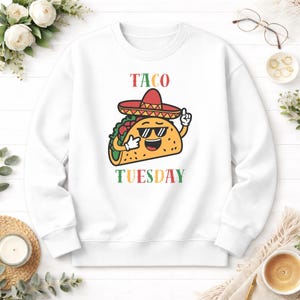May include: White sweatshirt with a cartoon taco wearing sunglasses and a sombrero. The text "TACO TUESDAY" is above and below the taco graphic. The taco is yellow with green, red, and brown details.