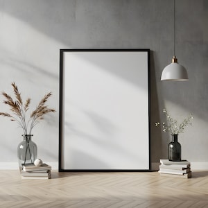 May include: A large, blank black framed poster leans against a gray wall on a wooden floor. A clear glass vase with dried plants sits on a stack of books to the left. A pendant light hangs from the ceiling, and a vase with greenery sits on books to the right.
