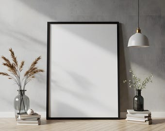 May include: A large, blank black framed poster leans against a gray wall on a wooden floor. A clear glass vase with dried plants sits on a stack of books to the left. A pendant light hangs from the ceiling, and a vase with greenery sits on books to the right.