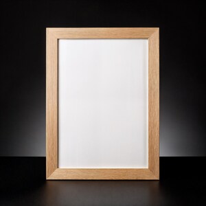 Könnte beinhalten: Ein rechteckiger Bilderrahmen aus Holz mit hellbraunem Finish. Der Rahmen hat einen weißen Innenraum und ist vor einem schwarzen Hintergrund platziert. Der Rahmen ist leer und bereit für ein Foto oder Kunstwerk.