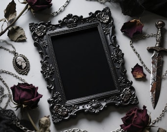 May include: A black ornate picture frame with a blank center, surrounded by dried burgundy roses, a silver chain, and a decorative dagger. A cameo pendant is also present, creating a gothic aesthetic.