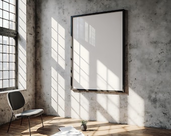 May include: An interior shot of a room with a large blank framed canvas on a concrete wall. Sunlight streams through a large window, casting shadows on the wooden floor. A gray chair, plant, and papers are in the room.