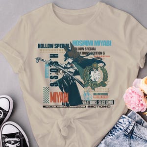 May include: A beige t-shirt with a graphic print of a character from the anime "Hoshimi Miyabi". The graphic features the character holding a sword and the text "H.S.O.S.G. Miyabi" and "Hollow Special Operations Section 6".
