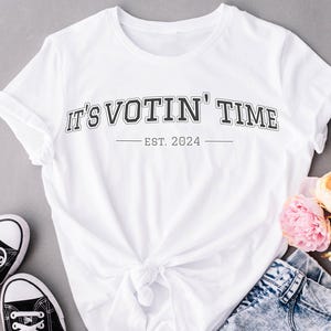 May include: White t-shirt with the text "IT'S VOTIN' TIME" in black, arched lettering, with "EST. 2024" below. The shirt is styled with black sneakers, denim shorts, and pink and peach flowers.