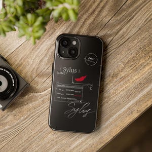 Op de afbeelding: Zwarte telefoonhoes met een grafisch ontwerp met de naam "Sylus", een rode veer en andere tekst. De hoes heeft een glanzende afwerking en ligt op een houten oppervlak.