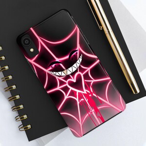 May include: A black phone case with a pink spiderweb design. The spiderweb has a pink heart in the centre and a pink grinning face with sharp teeth.