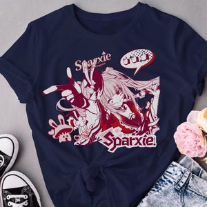 Pode incluir: Camiseta azul marinho com um gráfico em estilo anime vermelho e branco, com a palavra "Sparxie" e um balão de fala com polegares para cima. O design inclui uma ilustração de personagem. Tênis pretos e flores no fundo.