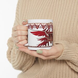 May include: White ceramic mug featuring a red and gold design of a stylised bird with outstretched wings. The design incorporates a red and gold cage with a gold crown on top.