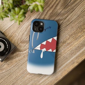 May include: A blue and white phone case with a shark's mouth design. The shark's teeth are white and the inside of the mouth is red. There is a white drip coming from the mouth.