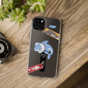 Peut inclure: Coque de téléphone transparente avec un motif de requin. L'étui présente un requin de dessin animé portant un chapeau blanc et une étiquette rouge avec un motif d'arête de poisson. Le mot "POP" est imprimé en haut de l'étui.