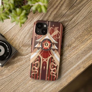 Puede incluir: Una funda para teléfono roja, blanca y dorada con un diseño geométrico. El diseño presenta un ojo grande con una estrella en el centro, rodeado de serpientes y una luna creciente.