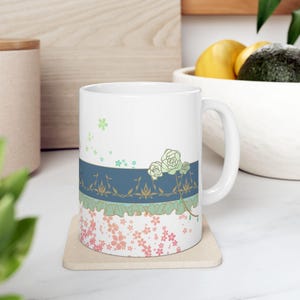 May include: White ceramic mug with a blue, green, and gold floral design. The design features a cluster of white roses with green leaves and stems. The mug has a white handle and is sitting on a beige coaster.