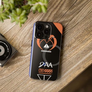 Peut inclure: Coque de téléphone noire avec un motif géométrique orange et blanc. La coque a un découpage pour l'objectif de l'appareil photo et le texte "DAA GB" imprimé dessus.