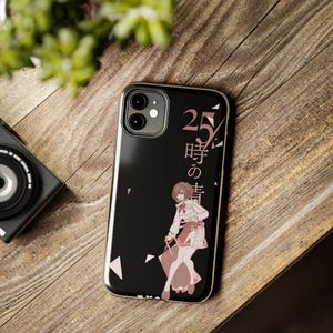 May include: Black phone case featuring a manga-style illustration of a character in a white and pink outfit, with Japanese text. The case has a clear border and is set on a wooden surface.