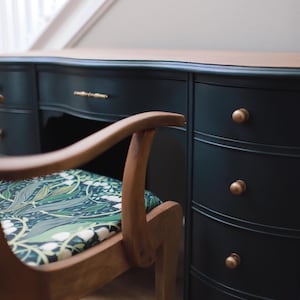 May include: A dark green desk with brass hardware and a wooden chair with a floral patterned cushion. The desk has multiple drawers and a curved front.