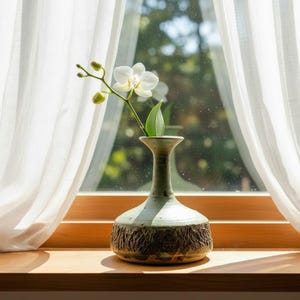 May include: A ceramic vase with a light green glaze holds a white orchid stem with a single leaf. The vase has a textured, brown band around its base. The vase sits on a wooden windowsill in front of a window with white curtains.