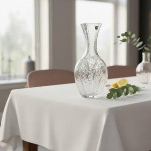 May include: A clear glass vase with a textured surface, on a white tablecloth. The vase has a slender neck and a wider base. A sprig of greenery and a lemon are on the table.