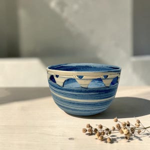 May include: A blue and white ceramic bowl with a distinctive design. The bowl has a striped pattern and a decorative rim with cut-out shapes. The bowl is on a light-coloured wooden surface.