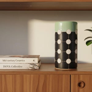 May include: A cylindrical ceramic vase featuring a matte black body with a grid of white circular dots and a solid light green top section. The vase stands upright, displaying a clean, geometric pattern across its exterior surface.