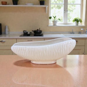 May include: A white ceramic serving bowl with a textured, organic design. The bowl has a wide, oval shape and sits on a small, round base. The background shows a kitchen setting with a window and shelves.