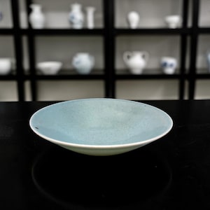 May include: A shallow, round, light blue ceramic bowl with a speckled texture. The bowl has a white rim and sits on a black surface. The background features a display of white and blue ceramic pieces on shelves.
