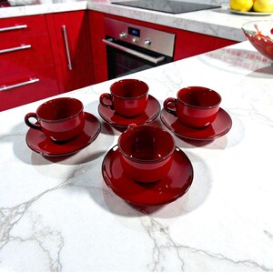 May include: Four red espresso cups and saucers on a white marble surface. Each cup has a handle and sits on a matching saucer. The cups and saucers have a dark trim. The kitchen has red cabinets.