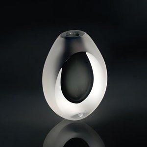 May include: A frosted glass vase with a silver-coloured, oval-shaped opening. The vase has a smooth, rounded shape and a small logo on the bottom.