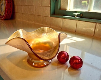May include: An iridescent amber glass bowl with a ruffled rim and etched floral design. Two small, round, red foil-wrapped candies sit beside the bowl on a white countertop. The bowl has a pedestal base.