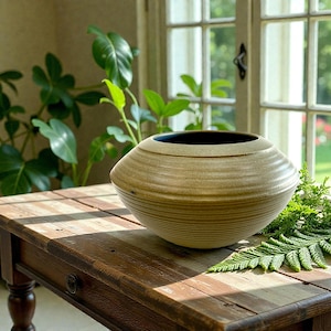 May include: A beige ceramic vase with a dark interior sits on a wooden table. The vase has a rounded, horizontal-striped design. The table is made of dark wood and is in front of a window with plants in the background.