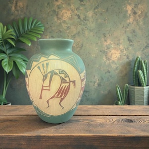 May include: A turquoise ceramic vase with a Kokopelli design. The vase features a cream-coloured central panel with a red and brown Kokopelli figure. The vase is on a wooden surface with plants in the background.