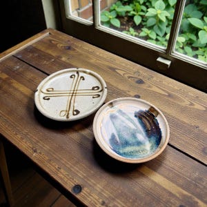 May include: Two handmade ceramic plates on a wooden table. One is oval with a cream base and brown cross design. The other is round with blue, brown, and white abstract patterns. The plates are near a window.