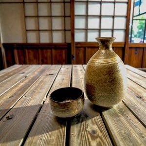 May include: A brown ceramic sake bottle and cup on a wooden table. The bottle has a rounded body and a narrow neck, with horizontal lines. The cup is small and round, with a dark interior. The table is made of wooden planks.