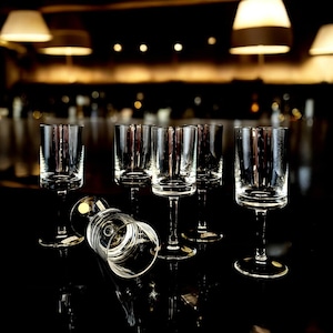 May include: A set of clear stemmed glasses with a short, cylindrical bowl. One glass lies on its side, while the others stand upright on a reflective black surface. The glasses are lit by overhead lighting, creating highlights and shadows.