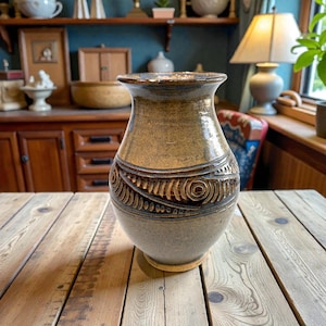 May include: A handcrafted ceramic vase with a wide base and flared rim. The vase has a textured, earthy brown glaze with a decorative band of carved patterns around the middle. The vase is displayed on a rustic wooden table.