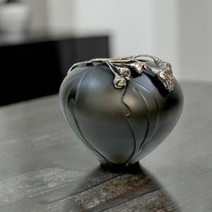 May include: A dark grey decorative vase with a silver floral design around the rim. The vase has a rounded shape with vertical, petal-like grooves. The vase is on a dark wooden surface.