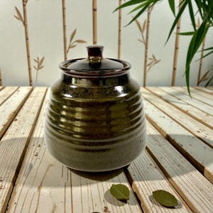 May include: A dark green ceramic jar with a lid. The jar has a rounded shape with horizontal grooves and a glossy finish. The lid is a darker shade of brown. The jar is likely used for storage and adds a rustic touch to any kitchen.