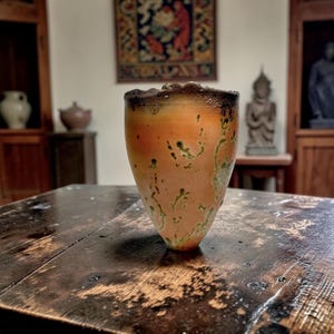 May include: A handmade ceramic vase with an organic shape. The vase has a warm orange base with green and brown accents, and a dark, textured rim. It is displayed on a dark wooden table, showcasing its rustic charm.