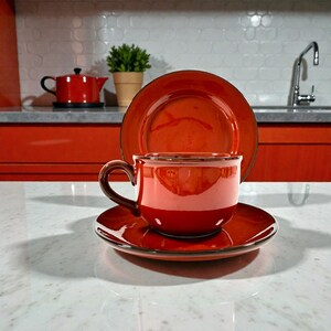 May include: A red ceramic teacup, saucer, and plate set. The cup has a brown handle and sits on a matching saucer. A plate is behind the cup. A red teapot is in the background. The set is on a white countertop.