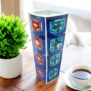 May include: A tall, blue and white ceramic vase with a geometric pattern of red, green, and gold accents. The vase is on a wooden table.