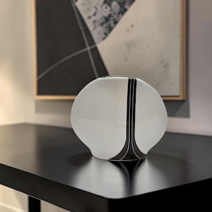 May include: A rounded, white glass vase featuring a distinct vertical black stripe pattern that tapers towards the base. The object has a smooth, opaque finish and a wide, bulbous body with a small circular opening at the top centre.
