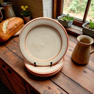 May include: A stack of cream-coloured ceramic plates featuring a thin, reddish-brown border. The plates are on a wooden surface, with a jug and a loaf of bread. The plates are held by a black plate stand.