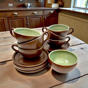May include: A collection of brown ceramic teacups and saucers with a pale green interior. The cups are stacked on a wooden table, with more cups and saucers in view. The overall style is rustic and inviting, ideal for a kitchen or dining area.